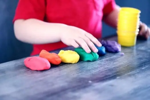 Vibrant modelling clay in red, orange, yellow, green, blue, and purple, with a child’s hand shaping the green clay on a dark wooden table, behind a stack of yellow bowls, promoting creative play and sensory activities.