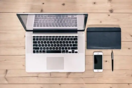 Laptop computer with open screen on wooden desk, alongside smartphone, notebook, and pen, representing productivity and remote work.