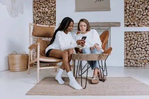 Young women sitting on stylish chairs and using a smartphone in a cozy, modern living room with wood log accents and contemporary decor.