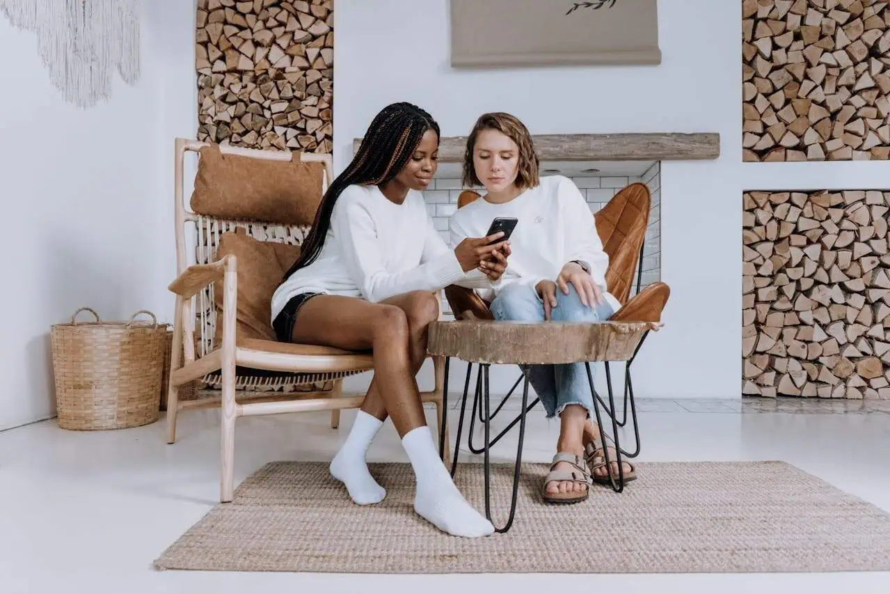 Young women sitting on stylish chairs and using a smartphone in a cozy, modern living room with wood log accents and contemporary decor.