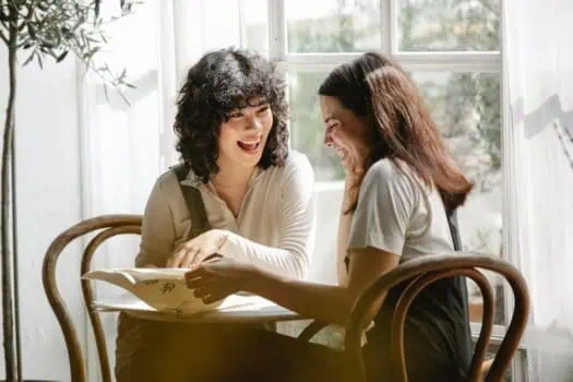 Happy women laughing and sharing a book at a coffee shop, cheerful friends enjoying conversation, warm natural light, cozy indoor setting, socialising, friendship, positive mood, community gathering, relaxed atmosphere, diverse women, modern cafe environment.