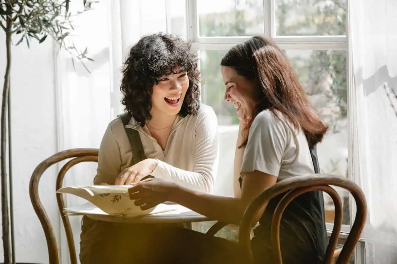 Happy women laughing and sharing a book at a coffee shop, cheerful friends enjoying conversation, warm natural light, cozy indoor setting, socialising, friendship, positive mood, community gathering, relaxed atmosphere, diverse women, modern cafe environment.