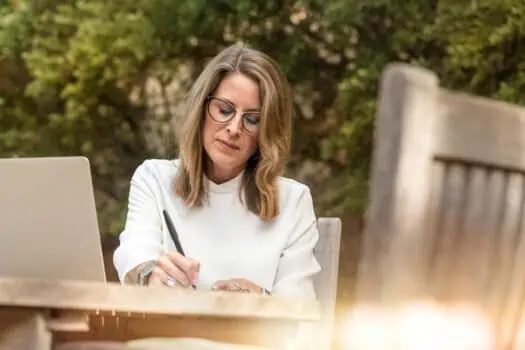 Thoughtful woman working outdoors with a laptop, writing in a notebook, representing focus, productivity, and outdoor work environments.