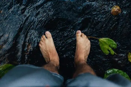 Bare feet standing in a shallow stream with floating leaves and water ripples, symbolising mindfulness, relaxation, and self-care - ideal for mental health and wellbeing themes.