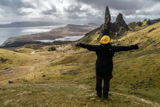 Vast Scottish Highlands landscape with a person in a black jacket and yellow beanie arms outstretched near iconic rock formations and rolling green hills, overlooking Loch and mountains, representing adventure and outdoor exploration.
