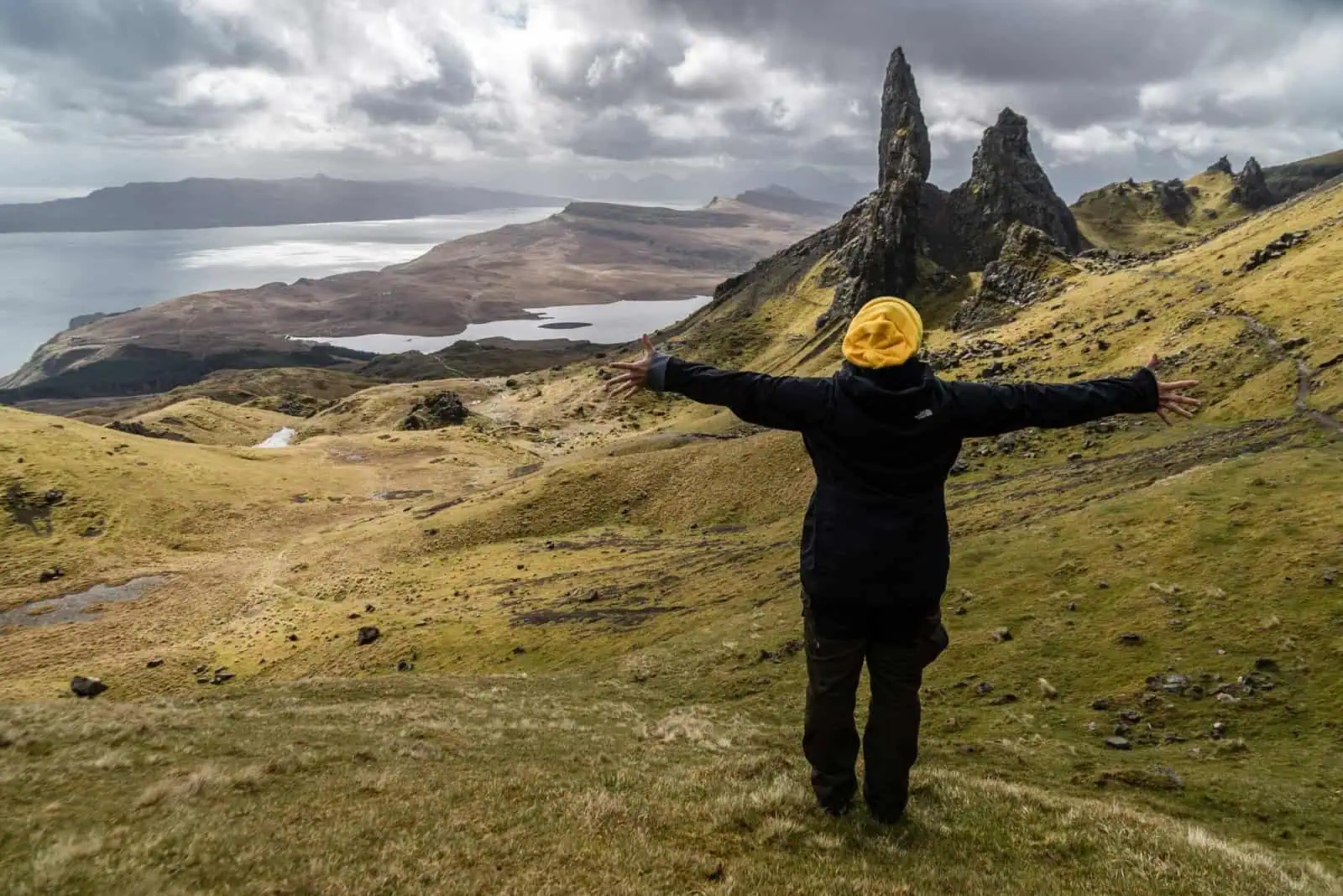 Vast Scottish Highlands landscape with a person in a black jacket and yellow beanie arms outstretched near iconic rock formations and rolling green hills, overlooking Loch and mountains, representing adventure and outdoor exploration.