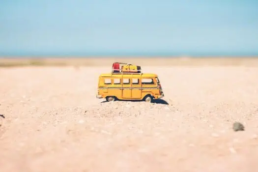 Yellow toy van with luggage on roof stranded on sandy beach under blue sky.