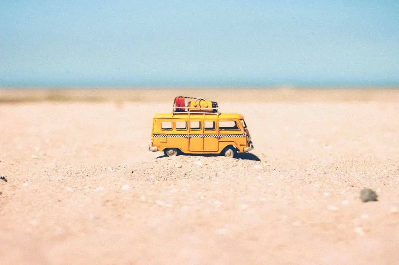 Yellow toy van with luggage on roof stranded on sandy beach under blue sky.