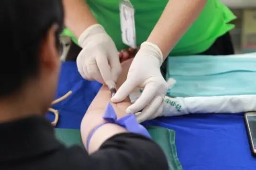 Needle being inserted into a volunteer's arm during a medical blood donation or blood test procedure, with healthcare professional wearing gloves.