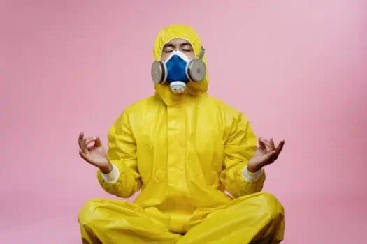 PPE health and safety professional meditating in protective yellow hazmat suit and respirator mask on pink background.