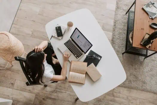 Laptop on white desk with open book, notebooks, and coffee; woman working at home office, studying, or practising skills; modern workspace with wooden and metal furniture.