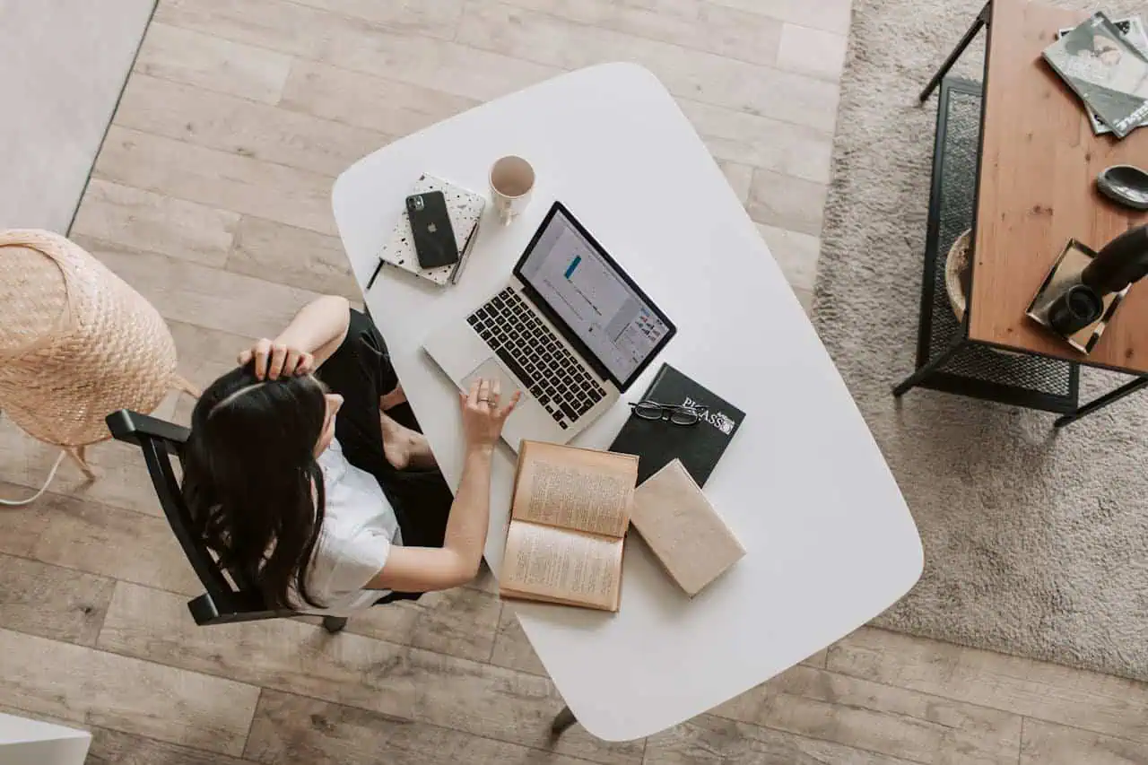Laptop on white desk with open book, notebooks, and coffee; woman working at home office, studying, or practising skills; modern workspace with wooden and metal furniture.
