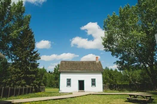 Small white wooden house with a sloped roof under a clear blue sky with white clouds, surrounded by green trees.