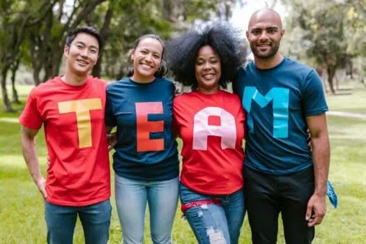 Bright diverse group of smiling young adults wearing colourful T-shirts spelling 'TEAM' outdoors in a park for teamwork and community spirit.