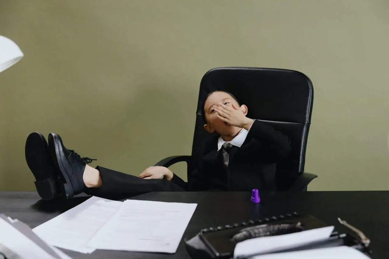 Child in business suit sitting with feet on desk, laughing and covering mouth, in a workplace setting with documents and office supplies, representing workplace humour or stress relief.
