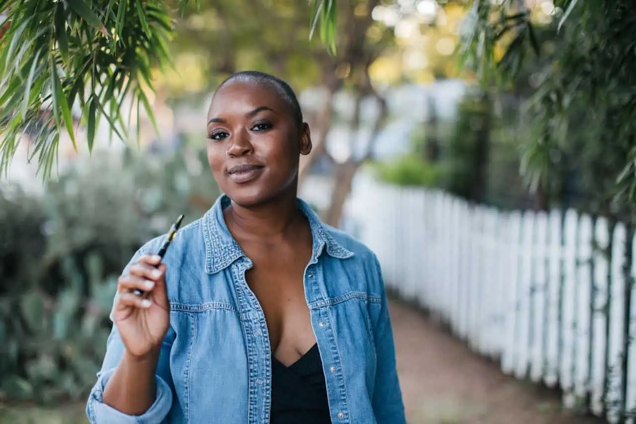 Confident young African woman smiling outdoors holding a pen, casual denim jacket, natural environment, empowering, professional, modern lifestyle, diverse community.