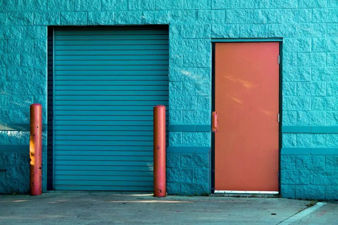 Garage door and pedestrian door exterior, blue brick wall, industrial building, safety bollards, urban environment, fire-resistant doors, commercial property, security access, loading dock, modern infrastructure.