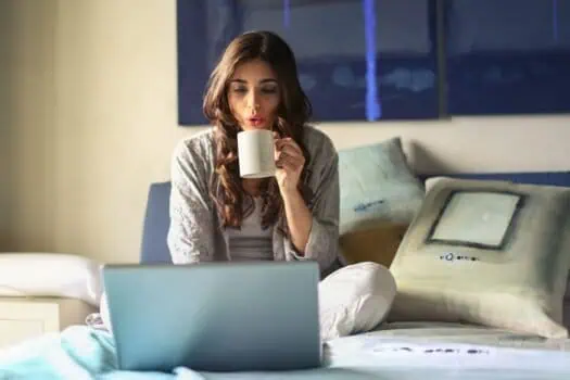 Relaxed woman working on laptop in bed, enjoying a hot drink at home, cozy bedroom setting, casual attire, morning or evening routine, modern lifestyle, comfort, digital work and relaxation.