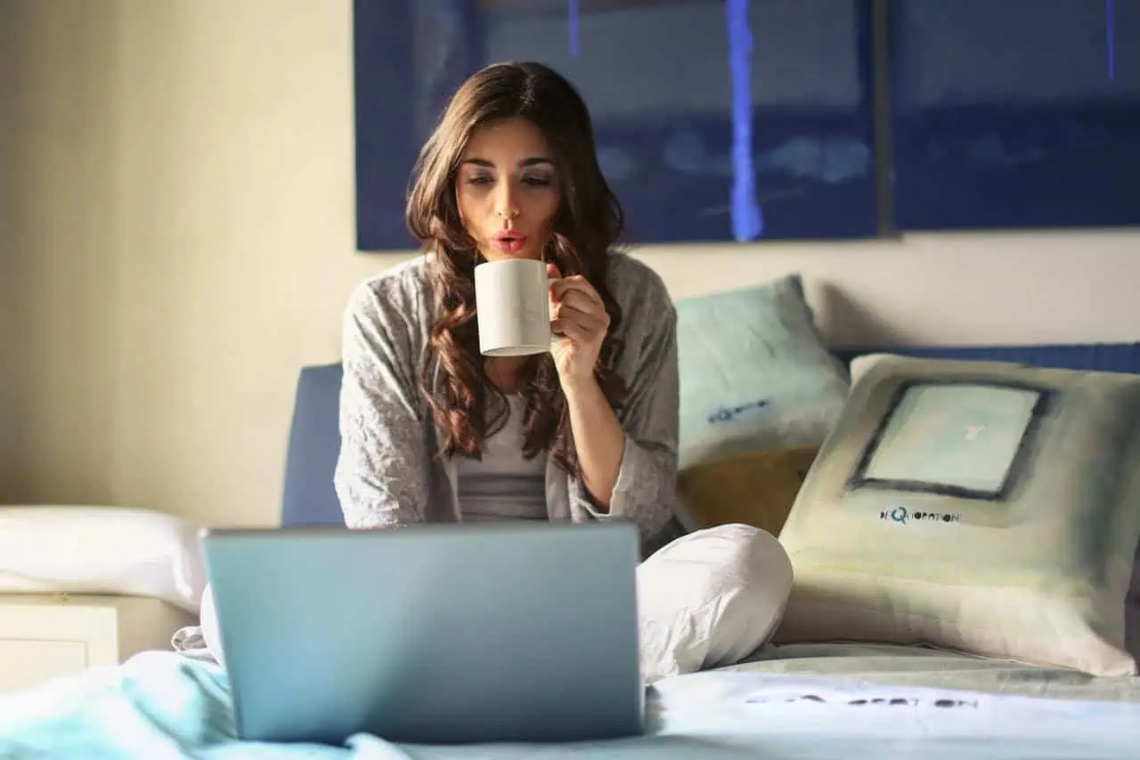 Relaxed woman working on laptop in bed, enjoying a hot drink at home, cozy bedroom setting, casual attire, morning or evening routine, modern lifestyle, comfort, digital work and relaxation.