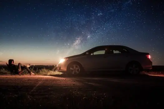 Starry night sky with a car parked on the roadside and a person relaxing in a camping chair, highlighting outdoor adventure and stargazing in the UK.