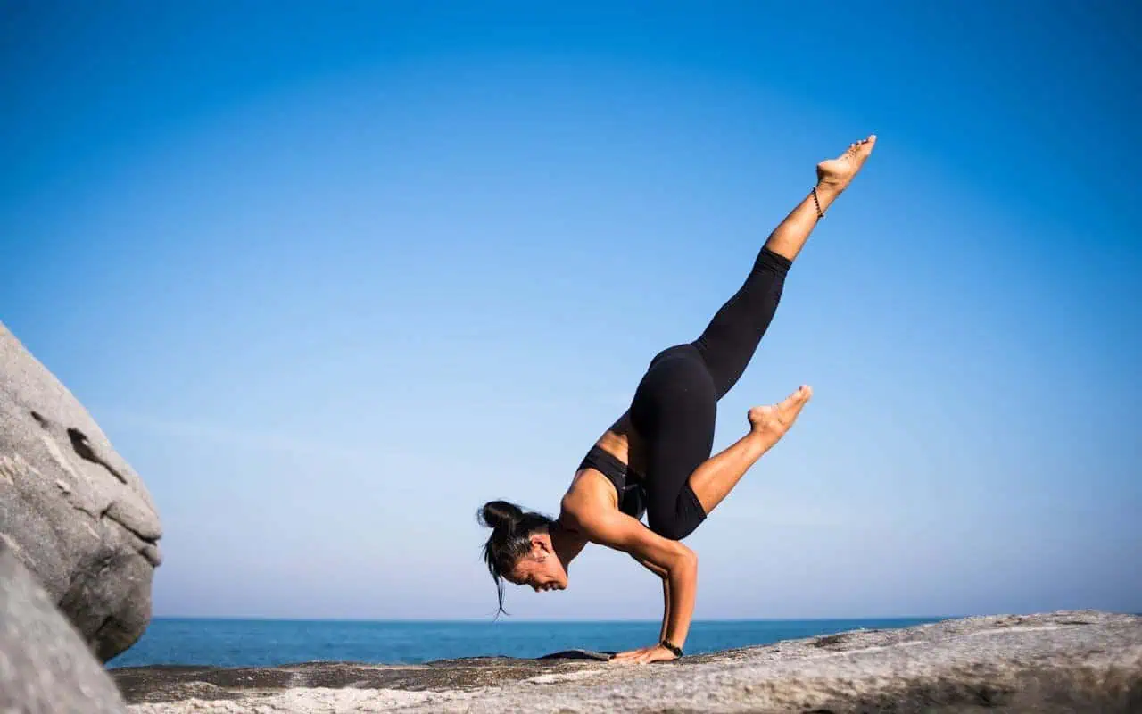 Handstand yoga pose on rocky seaside surface during daytime, woman balancing upside down with one leg extended up, practicing yoga mindfulness and flexibility by the ocean.