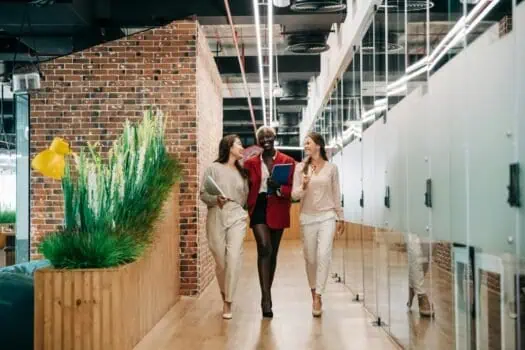 Conversational group of three diverse women walking and smiling in modern office corridor, holding notebooks and discussing, bright indoor workspace with exposed brick wall and glass lockers.