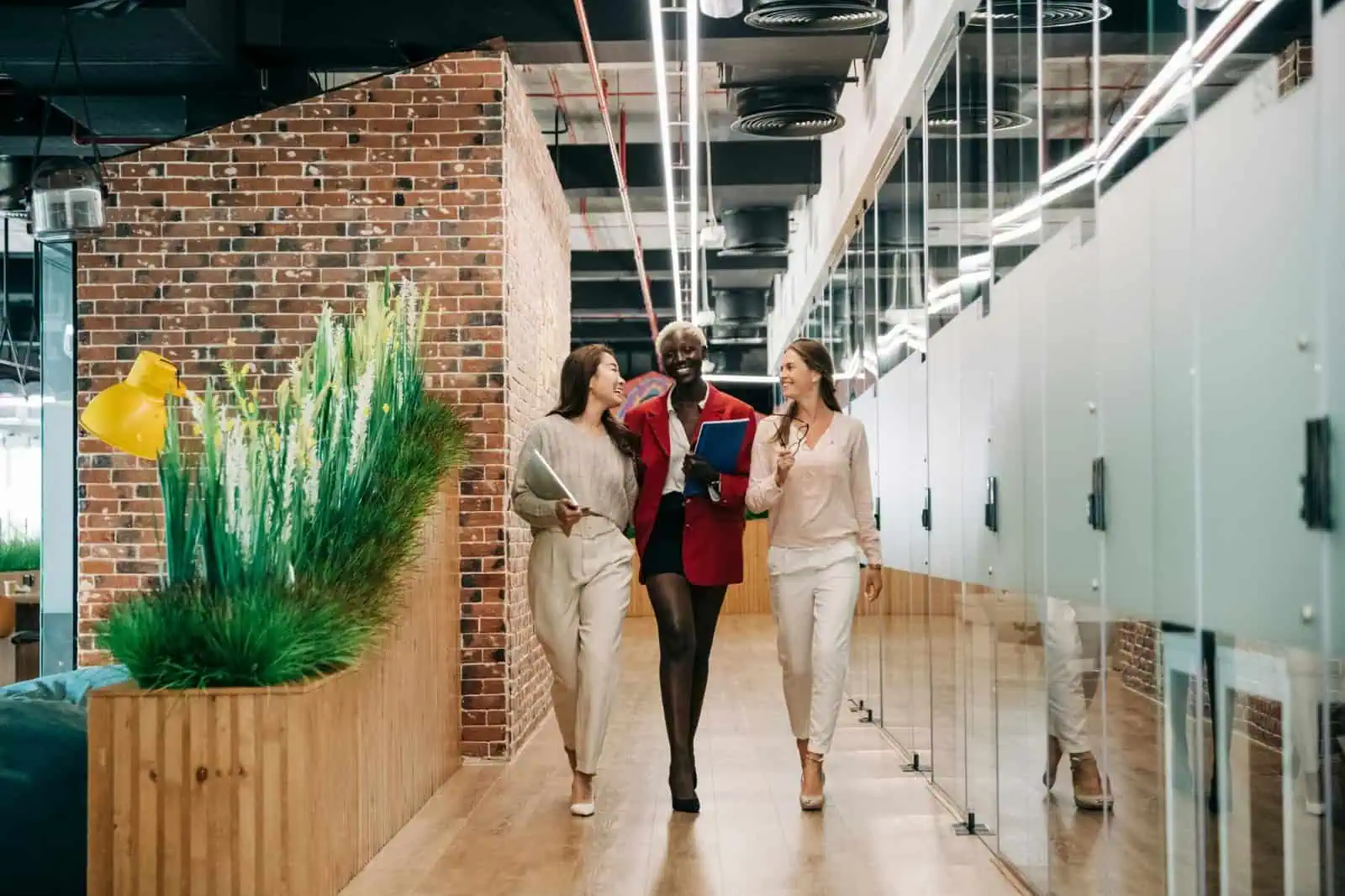 Conversational group of three diverse women walking and smiling in modern office corridor, holding notebooks and discussing, bright indoor workspace with exposed brick wall and glass lockers.