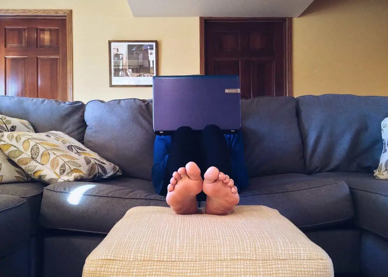 Laptop user with feet up on ottoman, sitting on a grey sofa at home.