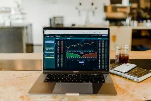 Laptop displaying financial trading charts on a wooden table with a glass of drink, iPhone, and a book in a modern workspace.