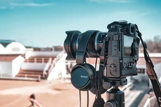High-quality mirrorless camera with professional-grade lens and Sony headphones, set outdoors on a tripod, ready for photography or videography. Captures a clear, bright sky with a modern urban backdrop.