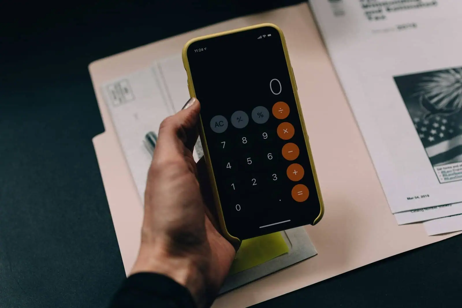 A person using a calculator app on a yellow smartphone, with documents and papers on a desk, illustrating finance and budgeting concepts.