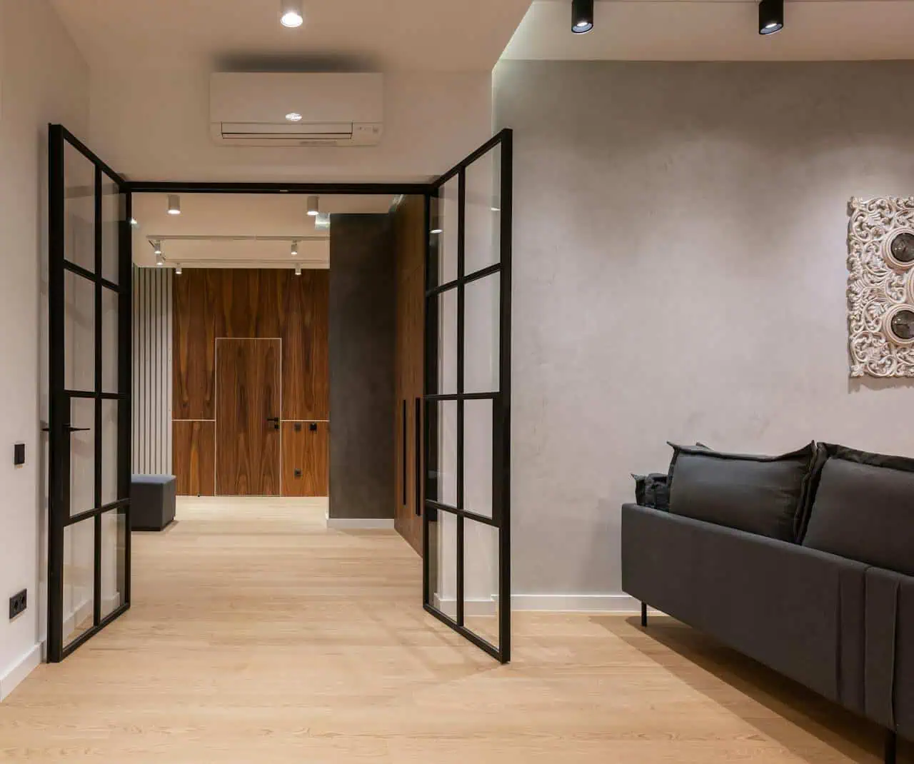 Modern living room with black-framed glass double doors, grey sofa, wooden flooring, contemporary wall art, and an air conditioning unit, showcasing stylish interior design.