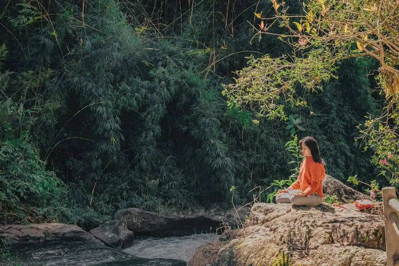 Meditating woman practicing mindfulness by river in lush forest, serenity and mental health, nature, relaxation, wellness, outdoor meditation, peaceful scene, stress relief, mental wellbeing, eco-friendly lifestyle, spiritual practice, Prowess SEO photo.