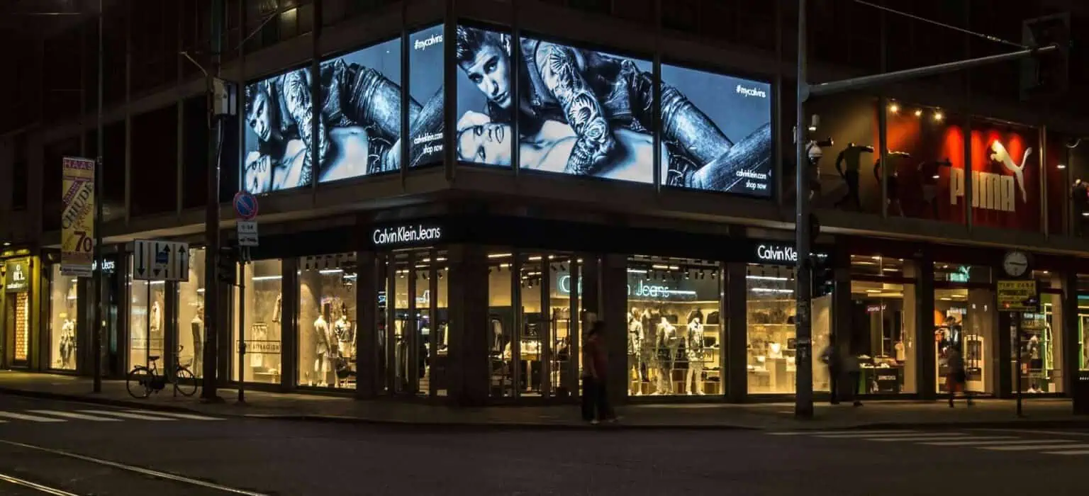 Brightly lit Calvin Klein Jeans store at night on a busy city street with large advertising screens displaying fashion models.