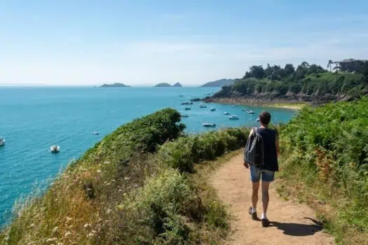 Beach trail with a person walking along a coastal path overlooking the sea and boats, sunny day, lush greenery, scenic seaside view, outdoor leisure activity, trekking, nature walk, coastal landscape.