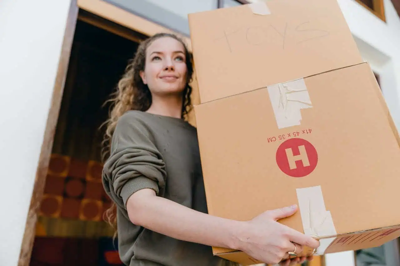 Large cardboard box with "TOYS" written on it being carried by smiling young woman moving house.