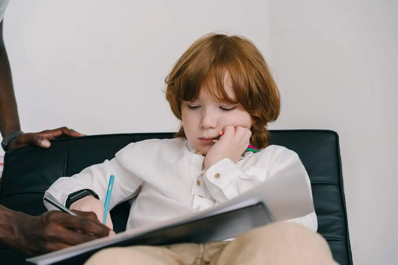 Frustrated young boy with red hair and freckles, sitting on a black sofa, holding a pencil and a notebook, feeling bored or disengaged in a home or classroom setting.