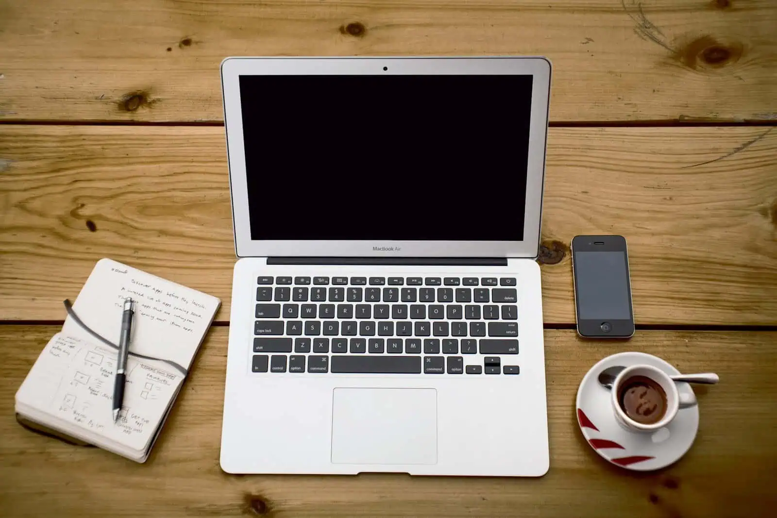 Laptop computer placed on a wooden desk with a smartphone, notepad with handwritten notes, pen, and a cup of coffee, illustrating workspace and productivity concept for Prowess.