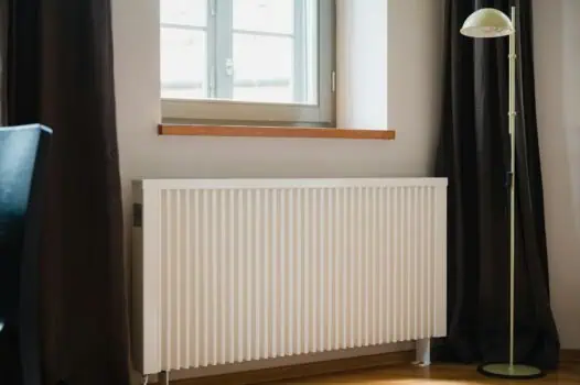 Bright interior room with a modern white radiator under a window, dark curtains, and a tall floor lamp, showcasing comfortable living space with natural sunlight and minimalist décor.
