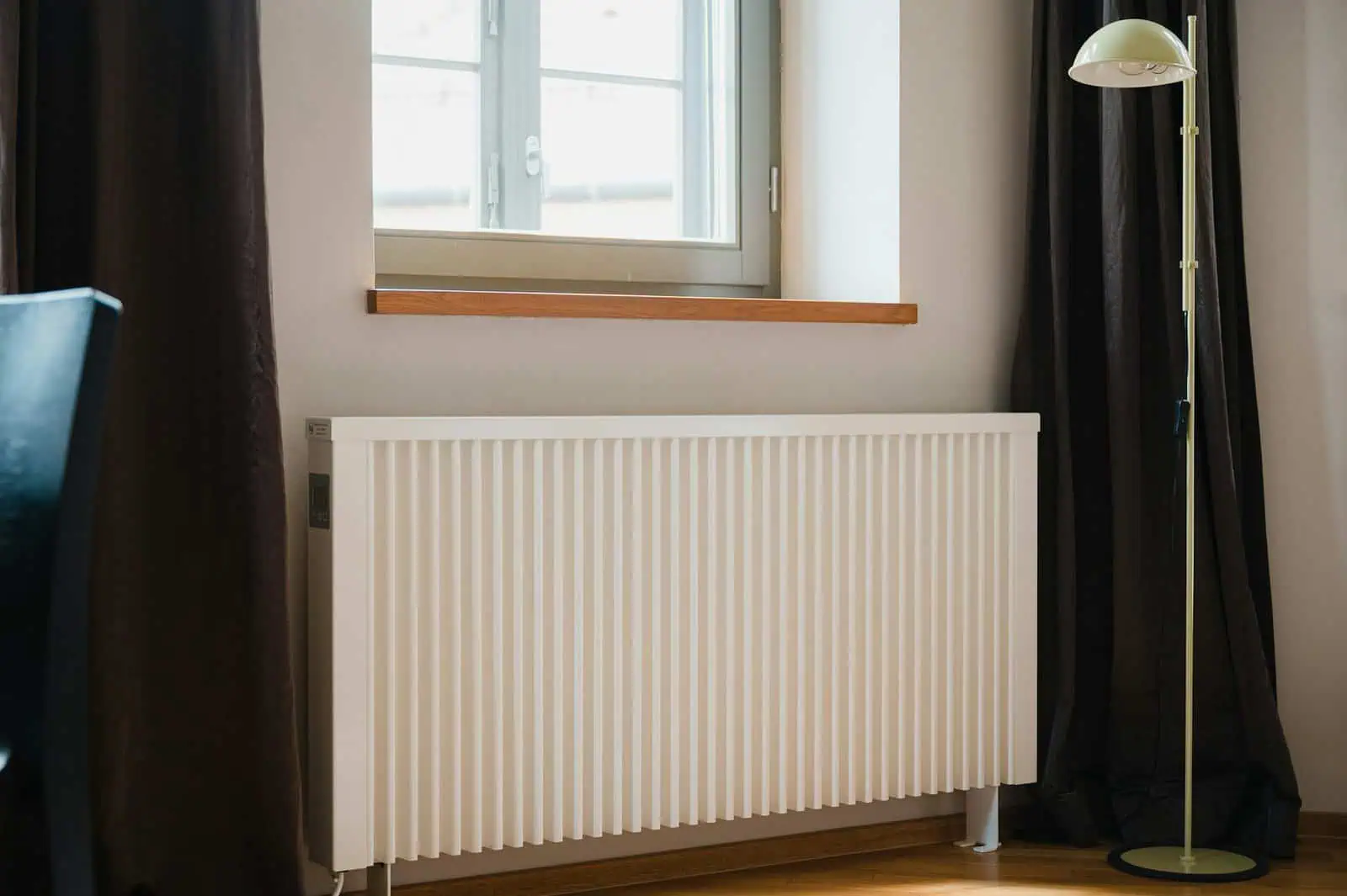 Bright interior room with a modern white radiator under a window, dark curtains, and a tall floor lamp, showcasing comfortable living space with natural sunlight and minimalist décor.