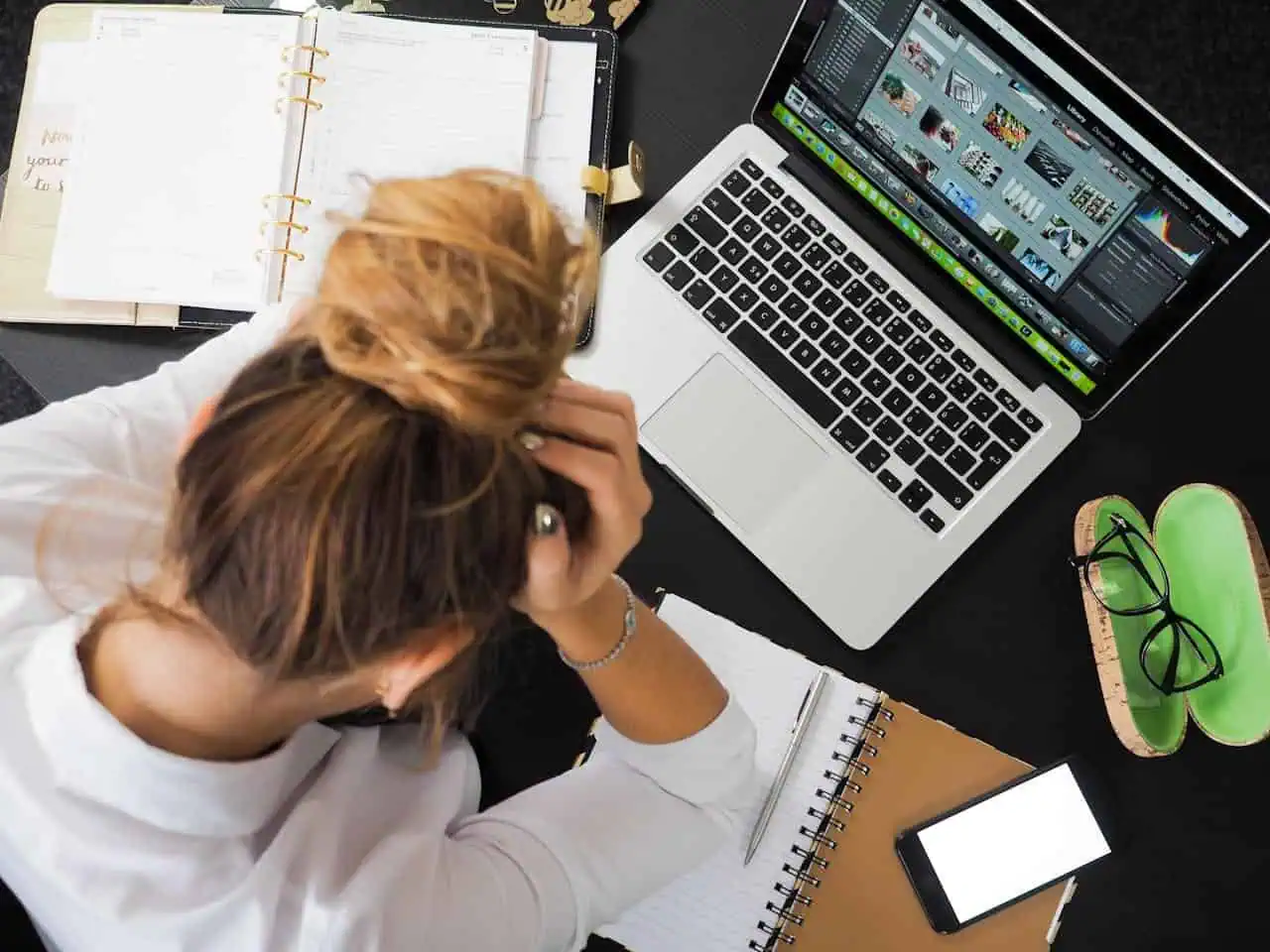 Overwhelmed person working on laptop with design software, surrounded by notebooks and accessories on a black desk.