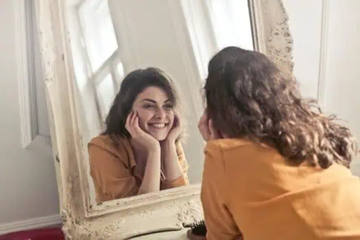 Bright young woman smiling at herself in a mirror, enjoying self-love and confidence, representing mental health and positivity.