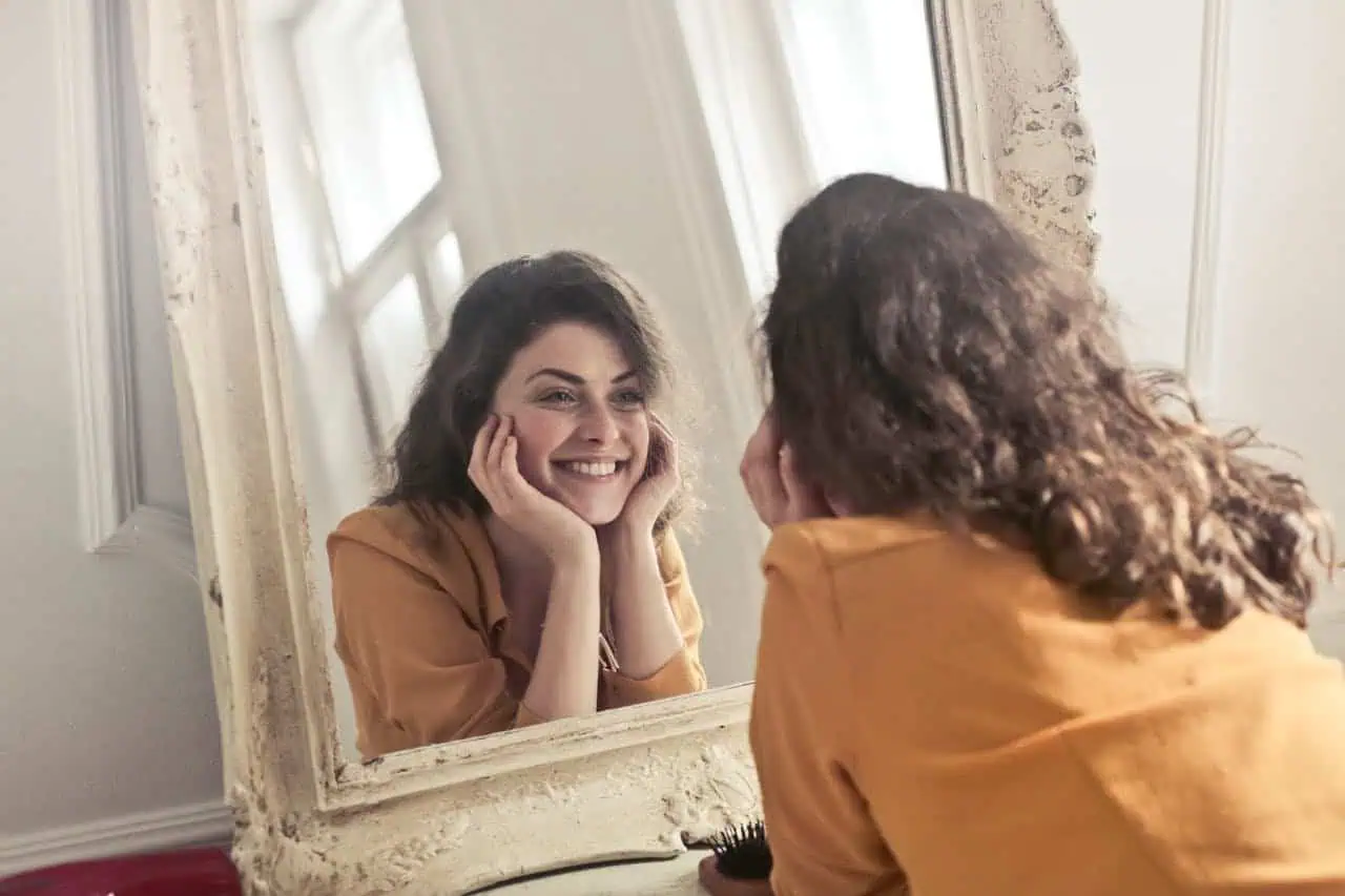Bright young woman smiling at herself in a mirror, enjoying self-love and confidence, representing mental health and positivity.