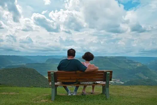 Bench with a view of green rolling hills and cloudy sky, couple sitting on a park bench enjoying nature and outdoor scenery, peaceful moment in countryside, scenic landscape, relaxing outdoors, nature appreciation, tranquility, community, wellbeing.