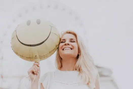 Bright woman holding a happy face balloon, celebrating joy and positivity, outdoors, representing mental health awareness and emotional well-being.