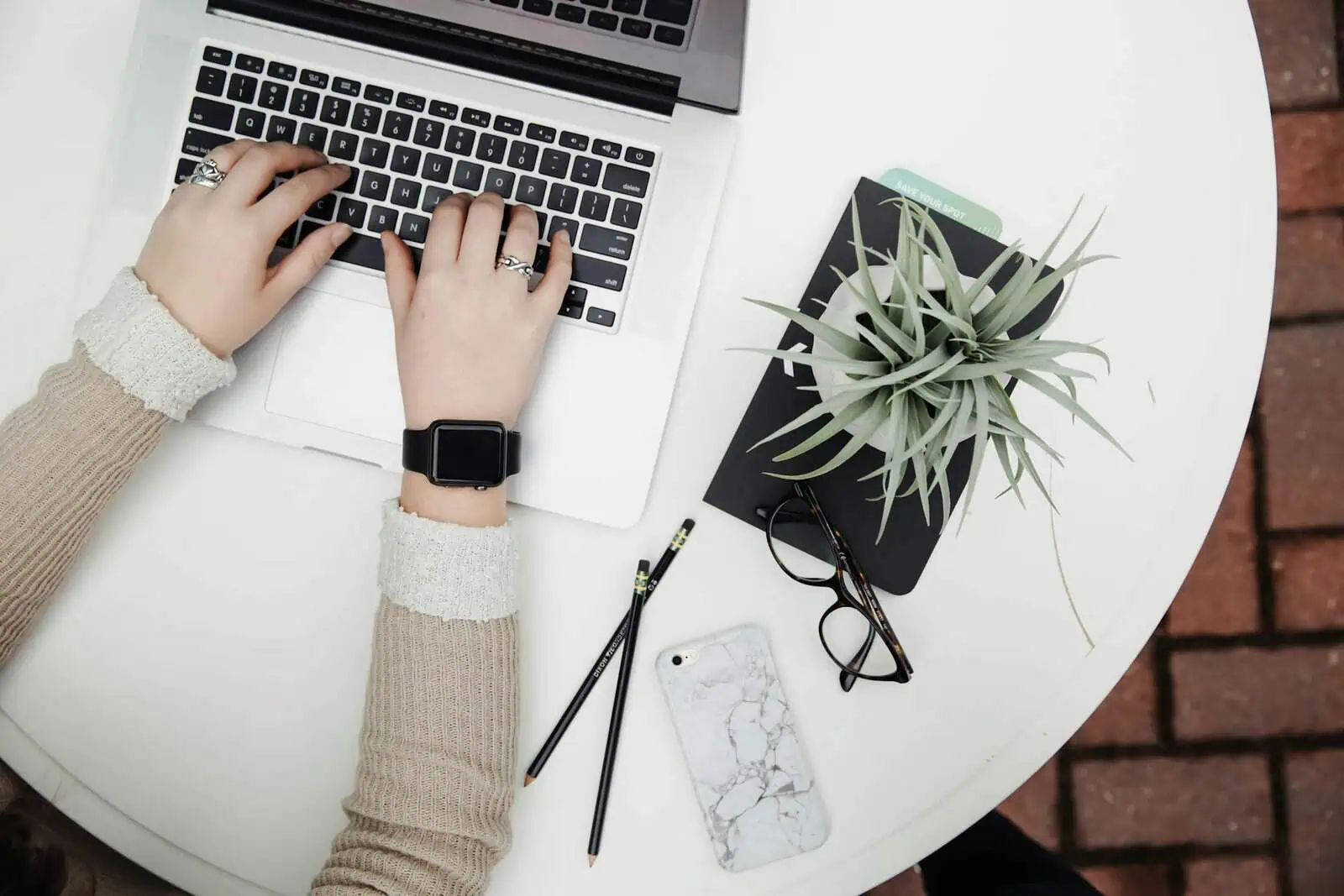 Laptop user working at a white desk with stationery, a potted plant, glasses, and a smartphone, capturing a modern, productive workspace with a focus on remote work and organisation.