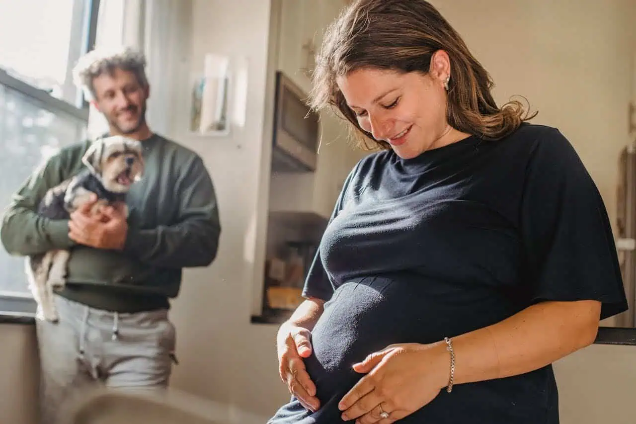 Smiling pregnant woman gently touching her belly with a man holding a playful puppy in the background inside a cosy home setting.