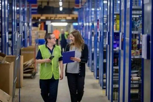 Staff and manager having a friendly discussion in a warehouse aisle filled with shelves and inventory, highlighting teamwork and logistics management for ecommerce or distribution centres.