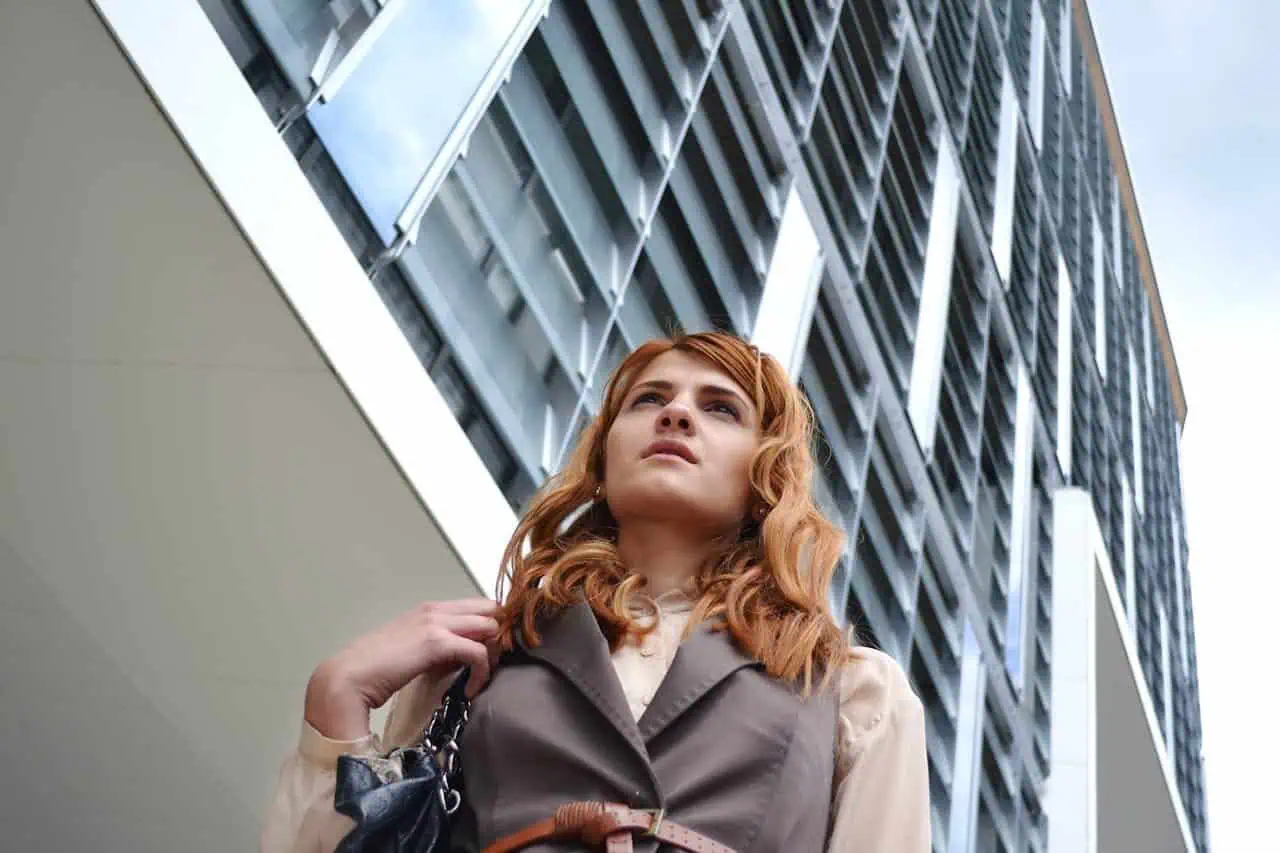 Urban woman with red hair looking up near modern building in a cityscape, professional business attire, outdoor shot, contemporary architecture, professional lifestyle.