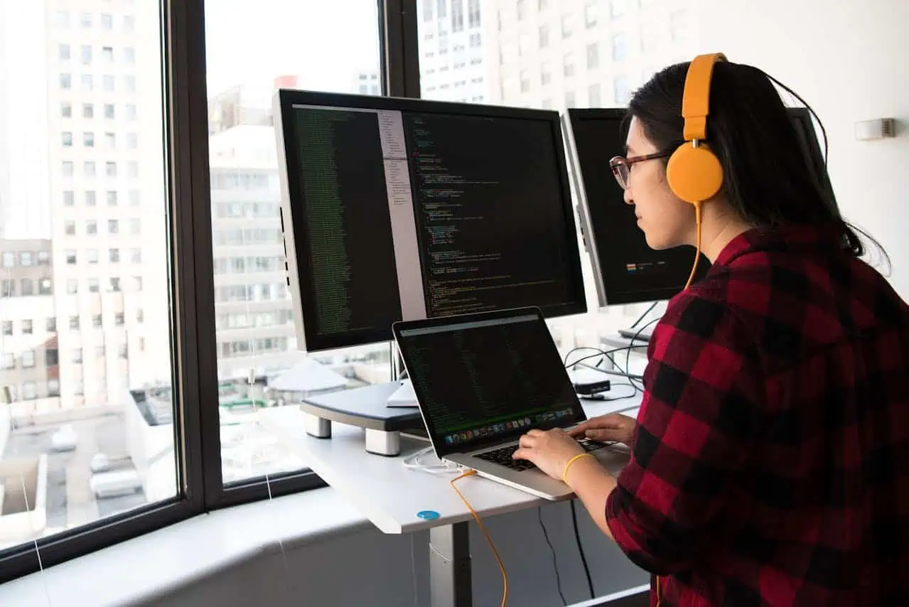 Coding professional woman working at laptop with multiple screens in bright office space.